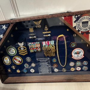 May include: A wooden display case with a blue interior, showcasing military medals, pins, and insignia. The case includes a U.S. Coast Guard emblem and a section with the American flag. A gold-colored necklace is also displayed.