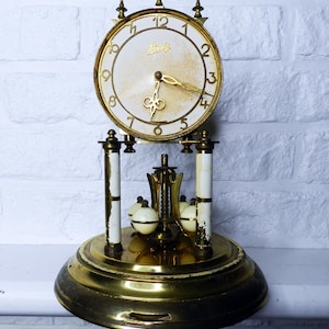 May include: An antique brass and white mantel clock with a round face. The clock face is white with gold numerals and hands. The clock has a decorative pendulum and is set on a brass base. The clock has the word "Lenzkirch" on the face.