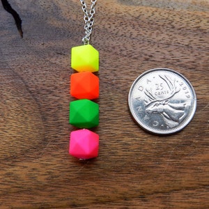 Puede incluir: Un collar de cadena plateada con cuatro cuentas cuadradas de color ne&oacute;n. Las cuentas son amarillas, naranjas, verdes y rosas. El collar est&aacute; sobre una superficie de madera junto a una moneda canadiense de 25 centavos.