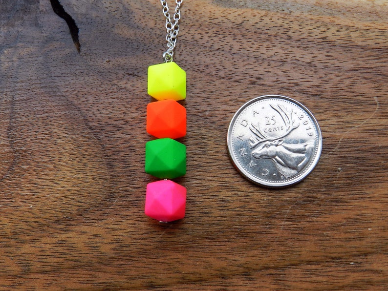 Puede incluir: Un collar de cadena plateada con cuatro cuentas cuadradas de color ne&oacute;n. Las cuentas son amarillas, naranjas, verdes y rosas. El collar est&aacute; sobre una superficie de madera junto a una moneda canadiense de 25 centavos.