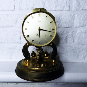 May include: A vintage brass clock with a cream-colored face and black hands. The clock features Roman numerals and a decorative base with three small brass spheres. The clock is set against a white brick wall.