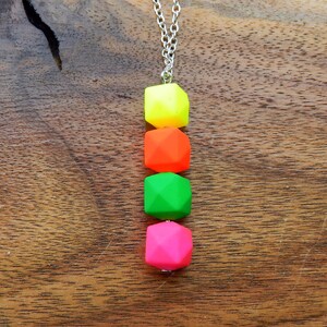 Puede incluir: Un collar de cadena plateada con cuatro cuentas geom&eacute;tricas de color ne&oacute;n. Las cuentas son amarillas, naranjas, verdes y rosas.
