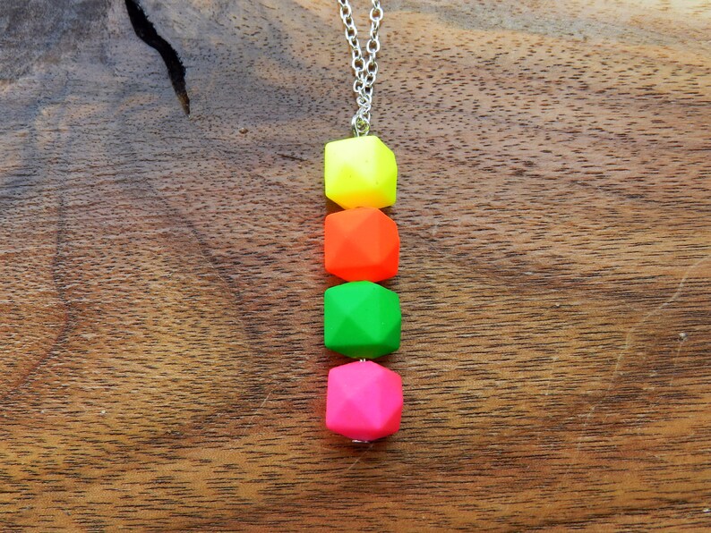 Puede incluir: Un collar de cadena plateada con cuatro cuentas geom&eacute;tricas de color ne&oacute;n. Las cuentas son amarillas, naranjas, verdes y rosas.