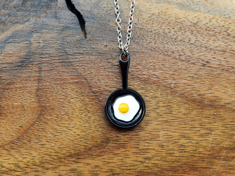 K&ouml;nnte beinhalten: Eine Halskette mit einer silbernen Kette und einem schwarzen Pfannenanh&auml;nger mit einem wei&szlig;en Spiegelei mit einem gelben Eigelb.