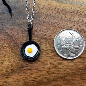 K&ouml;nnte beinhalten: Eine Halskette mit einer silbernen Kette und einem schwarzen Pfannenanh&auml;nger mit einem wei&szlig;en Spiegelei mit einem gelben Eigelb. Der Anh&auml;nger ist etwa so gro&szlig; wie eine kanadische 25-Cent-M&uuml;nze.