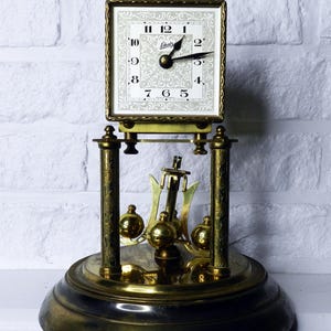 May include: A vintage brass-toned mantel clock with a square face and ornate detailing. The clock face is white with black numerals and hands, and the brand name "Schatz" is visible. The clock features a pendulum and a decorative base.