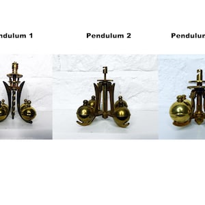 May include: Three antique brass clock pendulums are displayed against a white brick background. Each pendulum has a unique design, with spherical weights and ornate metalwork. The text "Pendulum 1", "Pendulum 2", and "Pendulum 3" are above each.