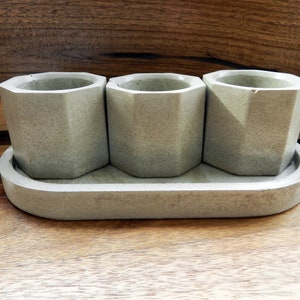 May include: Three small, grey concrete planters with a geometric design, sitting in a matching grey concrete tray.
