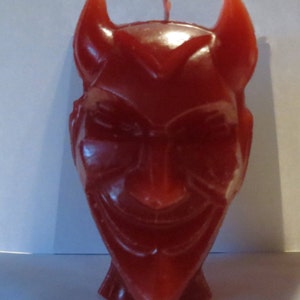 Puede incluir: Una vela de cera roja con forma de cabeza de diablo con cuernos. La vela tiene una cara detallada con una sonrisa traviesa.