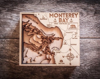 May include: A wooden map of Monterey Bay, California, with a compass rose and the text "Monterey Bay Area - 449 Miles". The map is carved with details of the coastline and surrounding areas, including Monterey Canyon, Pacific Grove, Carmel, and Big Sur.
