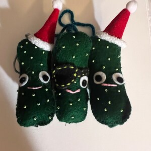 May include: Three green felt pickle ornaments with white felt trim and red felt Santa hats. The middle pickle has a black felt eye patch.