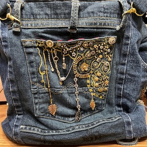 Könnte beinhalten: Eine Jeans-Tasche mit einer großen Tasche auf der Vorderseite. Die Tasche ist mit goldenen und silbernen Verzierungen verziert, darunter Ketten, Perlen und Anhänger. Die Tasche hat einen goldenen Verschluss und einen Riemen.