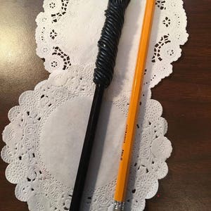 May include: A black and green wand with a green glass orb on top, lying on a white doily next to a yellow pencil with the text "AUTOMATIC No. 2" printed on it.