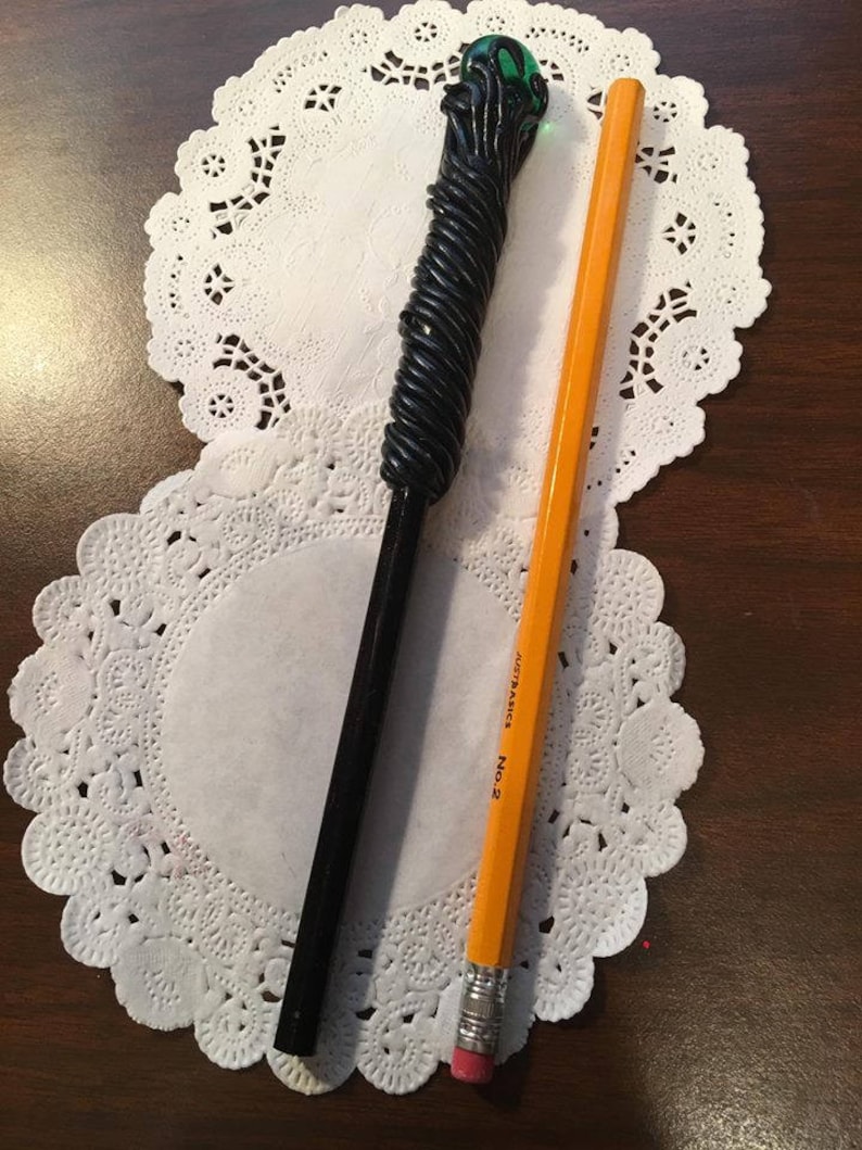 May include: A black and green wand with a green glass orb on top, lying on a white doily next to a yellow pencil with the text "AUTOMATIC No. 2" printed on it.