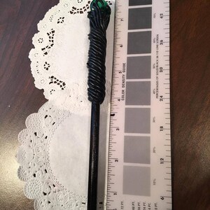 May include: A black and green wand with a twisted design and a green glass orb at the top. The wand is about 7 inches long and is measured against a ruler.