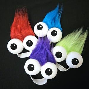 Puede incluir: Cuatro diademas peludas de colores vivos con grandes ojos saltones blancos. Las diademas son roja, azul, verde y morada.
