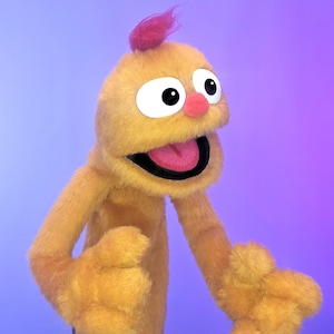 Puede incluir: Una marioneta de mano de color amarillo dorado y textura difusa, con una cabeza redondeada y un pequeño mechón de pelo rosa en la parte superior. Tiene ojos grandes blancos con pupilas negras, una nariz pequeña rosa y la boca abierta.