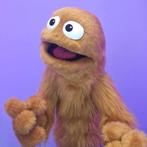 Può includere: Un burattino arancione e peloso con grandi occhi bordati di bianco e una bocca spalancata che rivela una lingua rosa. Il burattino ha una consistenza ruvida e braccia tese, su uno sfondo viola.