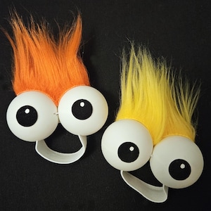 May include: Two fuzzy toy characters with large white googly eyes and elastic bands. One character has orange fur and the other has yellow fur.
