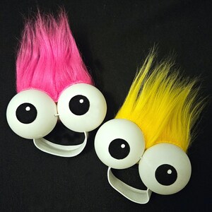 May include: Two fuzzy toy characters with large white eyes and black pupils. One character has pink fur and the other has yellow fur. Both characters have elastic bands around their heads.