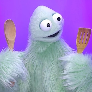 May include: A light green, furry puppet with large, googly eyes and a smiling expression. The puppet holds a wooden spoon in one hand and a wooden spatula in the other. The background is a gradient of purple and blue.