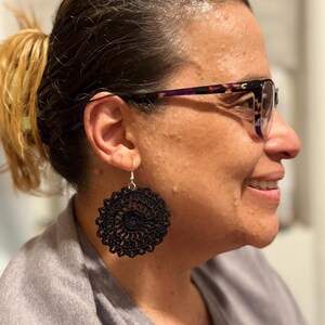 May include: A close-up of a black, crocheted earring with a circular design. The earring is suspended from a silver hook. The earring is worn by a person wearing glasses and a gray shirt.