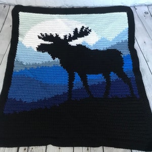 May include: A crocheted blanket with a silhouette of a moose standing in front of a mountain range. The mountains are a gradient of blue colors, and the background is black.