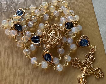 Puede incluir: Un rosario de oro con cuentas de vidrio blancas y azules. La pieza central presenta una imagen dorada de la Virgen María. El rosario está sobre un fondo dorado.