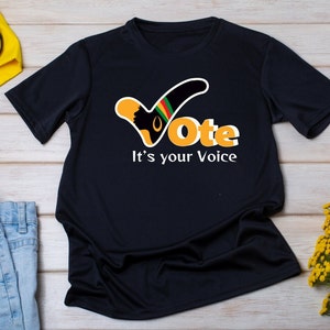May include: Black t-shirt with a graphic that says "Vote" in orange and white, with the words "It's your Voice" below. The graphic includes a silhouette of a person's profile and a checkmark design with green, yellow, and red stripes.