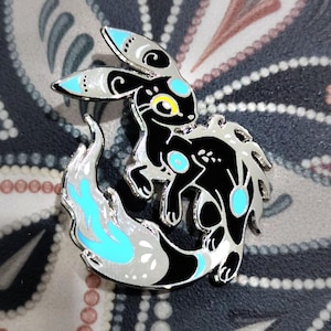 Puede incluir: Un pin de esmalte negro y plateado con una criatura estilizada con detalles azules y amarillos. El pin tiene una cola en forma de llama y está sobre un fondo estampado. El texto "© DRAGIBUZ / Lorène BARIOZ 2025" está en la parte inferior.