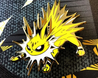 May include: Gold-colored enamel pin featuring a cartoon-style electric-type Pokemon with yellow and white fur, black eyes, and a white lightning bolt around its head.