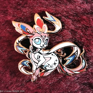 Puede incluir: Pin de esmalte dorado con una criatura mítica estilizada de color rosa y blanco con detalles azules y rojos. La criatura tiene una cola larga y fluida y una corona en la cabeza.