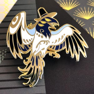 May include: A gold-colored metal pin featuring a stylized bird with a hat. The bird has white, black, and blue details. The pin has a shiny finish and is set against a patterned background. The bird's wings are spread, and it has multiple eyes.