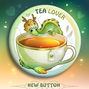 Puede incluir: Un dragón verde amante del té con una cola amarilla y verde está enroscado en una taza de té blanca con una bolsita de té. La taza de té está sobre un fondo azul claro con el texto "TEA LOVER" en un círculo blanco. El texto "NEW BUTTON & MAGNET" está debajo de la taza de té.