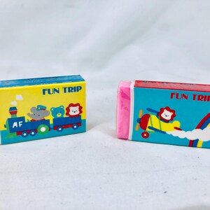 May include: Two rectangular erasers with colorful packaging. One features a train with animal characters and the text "FUN TRIP". The other shows an airplane with a lion and a rainbow, also with the text "FUN TRIP".