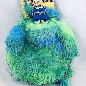 Peut inclure: Un porte-CD/DVD rond et duveteux, aux tons de bleu et de vert, ressemblant à un monstre. Il possède de grands yeux globuleux bleus et une étiquette jaune indiquant "12 CDs/DVDs" et "Baby Jungle". Le support peut contenir 12 CD ou DVD.