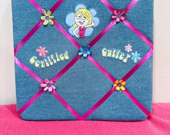 May include: A square blue denim memo board featuring a crisscross pattern of bright pink ribbon. The board is decorated with embroidered flower patches, a cartoon character patch, and the yellow embroidered text Certified Cutie.