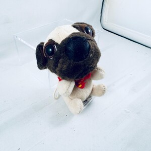 May include: A small plush pug toy featuring a cream-colored body, a dark brown face, and large black plastic eyes. The stuffed animal wears a small red collar with a yellow accent around its neck and has a white tag attached to its side.