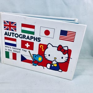 以下が含まれることがあります： さまざまな国旗の上に「AUTOGRAPHS」という文字が書かれた白いサイン帳。表紙にはハローキティとネズミの漫画の画像も表示されています。
