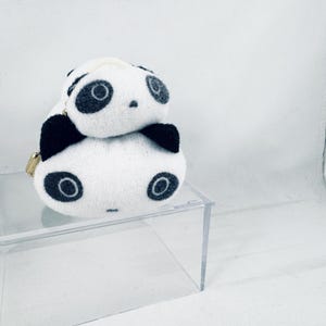 May include: A plush panda-shaped coin purse featuring a white body with black ears and dark grey circular eye patches. The soft, rounded accessory has a small gold-toned zip pull on the side and a secondary smaller panda head attached on top.