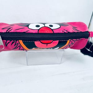 May include: A pink pencil case with a cartoon character design, featuring large white eyes, a red nose, and an open mouth. It has a black zip and a small black pom-pom on the zip pull. The case is cylindrical in shape.