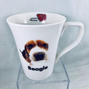 May include: White ceramic mug featuring a Beagle dog illustration. The mug has a curved handle and the words "Beagle" and "The Dog Artist Collection" are printed on the mug.