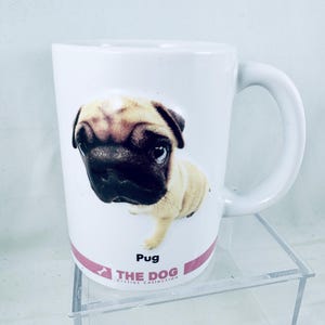 May include: A white ceramic mug featuring a high-angle, wide-lens photograph of a tan pug with a black face. Below the dog, a pink horizontal band displays the text "Pug", "THE DOG", and "Artlist Collection" with a small white bone icon.