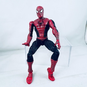 May include: A posable action figure of a superhero character featuring a red and black suit with a web-like pattern across the red sections. The figure has a black spider emblem on the chest, white triangular eyes, and articulated joints.