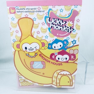 May include: A rectangular card featuring the text Lucky Monkey Banana House. The design includes a large yellow banana, a yellow monkey, a blue monkey, and a pink monkey against a background of small yellow bananas and blue stars.
