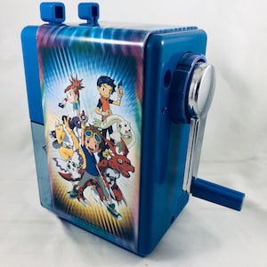 Peut inclure: Taille-crayon bleu avec un graphisme coloré de personnages d'anime. Le taille-crayon a une manivelle argentée et deux trous d'entrée pour les crayons. Le graphisme présente un fond tie-dye.