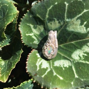 Puede incluir: Un colgante de alambre plateado envuelto alrededor de una piedra de ópalo blanco. El colgante se muestra en una hoja verde con marcas blancas.