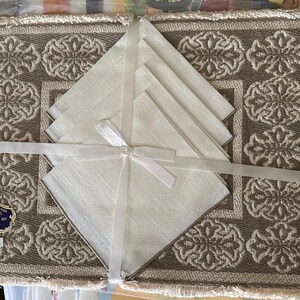 May include: A luncheon set featuring a beige placemat and napkins, tied with a white ribbon. The placemat has a floral pattern and fringe trim. The napkins are folded and stacked. The set includes a tag that reads "Kinsdale Creation".