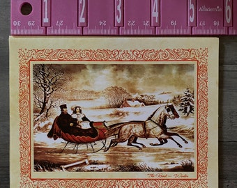 May include: A vintage winter scene print featuring a horse-drawn sleigh with two passengers. The sepia-toned image is framed with a red and cream border. The text "The Road - Winter" is at the bottom. The print measures approximately 5 inches wide.
