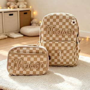 Peut inclure: Un sac à dos et un sac à déjeuner rectangulaire assorti à carreaux beiges et blancs. Les deux articles portent le nom Mason en lettres en relief texturées de couleur beige. Le sac à dos comporte une petite fleur crochetée blanche sur le devant.